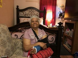 Elderly Hispanic woman in a pink shirt sitting on a chair with a broken arm and blue cast in her bedroom.